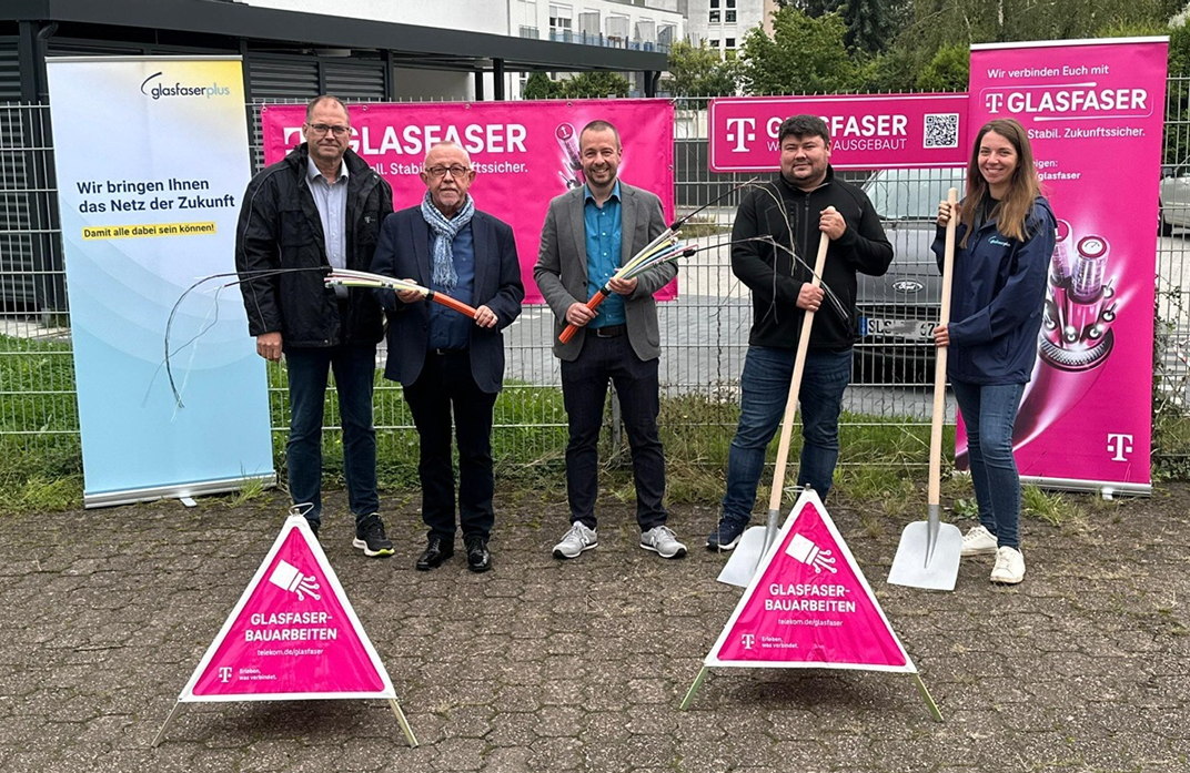 Fünf Personen stehen im Freien vor Bannern, die für den Glasfaser-Ausbau in Deutschland werben, halten Bauwerkzeuge und Kabel in der Hand, während auf dem Boden Schilder über Glasfaser-Installationsarbeiten angebracht sind.