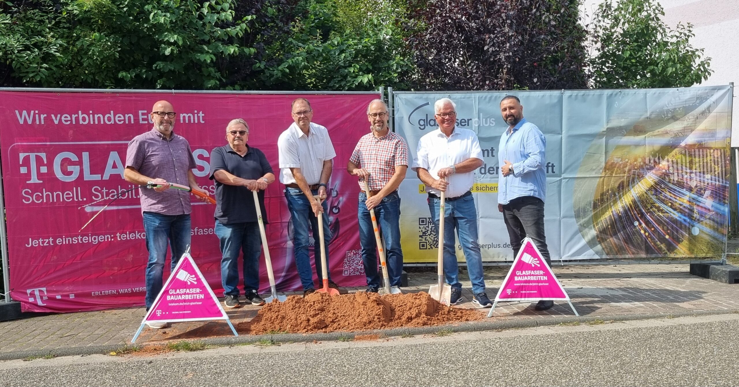 Sechs Männer stehen mit Schaufeln bei einem Spatenstich für den GlasfaserPlus-Glasfaserausbau. Hinter ihnen sind Markenschilder und Banner zu sehen, die auf den Glasfaser-Ausbau in Deutschland hinweisen.