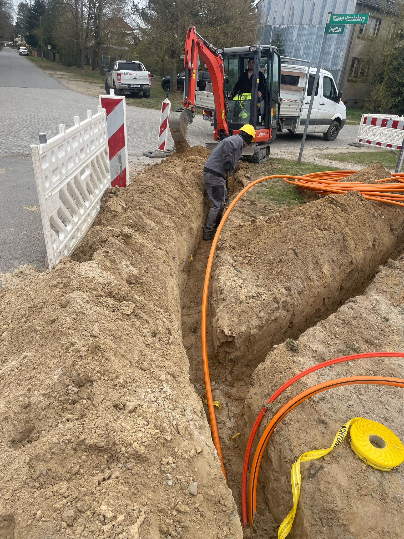 Ein Arbeiter mit Schutzhelm steht in einem Graben neben orangefarbenen Versorgungskabeln, während im Hintergrund ein Bagger und Fahrzeuge auf einer Glasfaser-Ausbau-Baustelle am Straßenrand zu sehen sind.