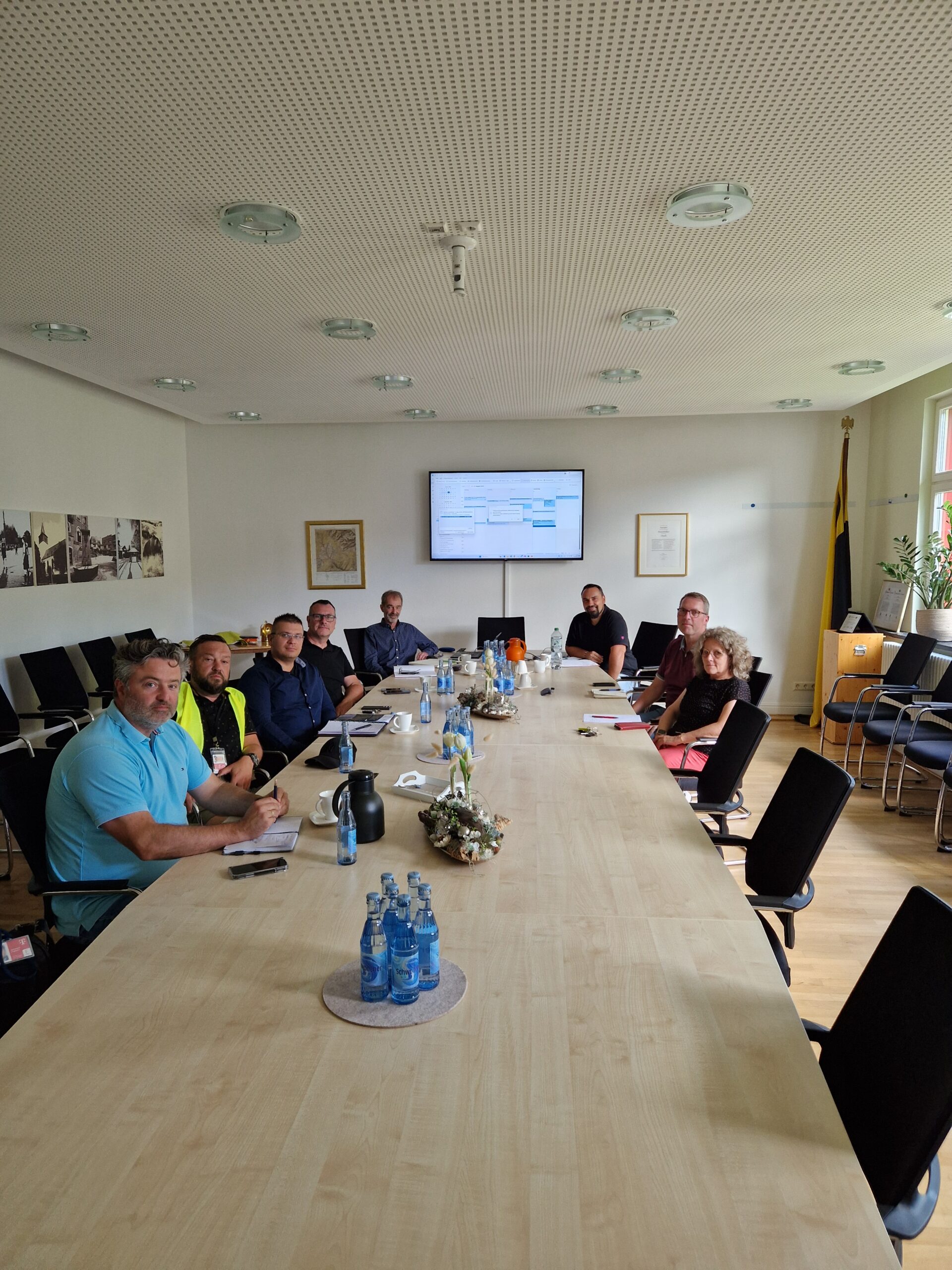 Neun Personen sitzen um einen langen Konferenztisch mit Getränken und Papieren, den Blick in die Kamera gerichtet. Am anderen Ende des Raumes zeigt ein Bildschirm Informationen über Glasfaser-Ausbau.