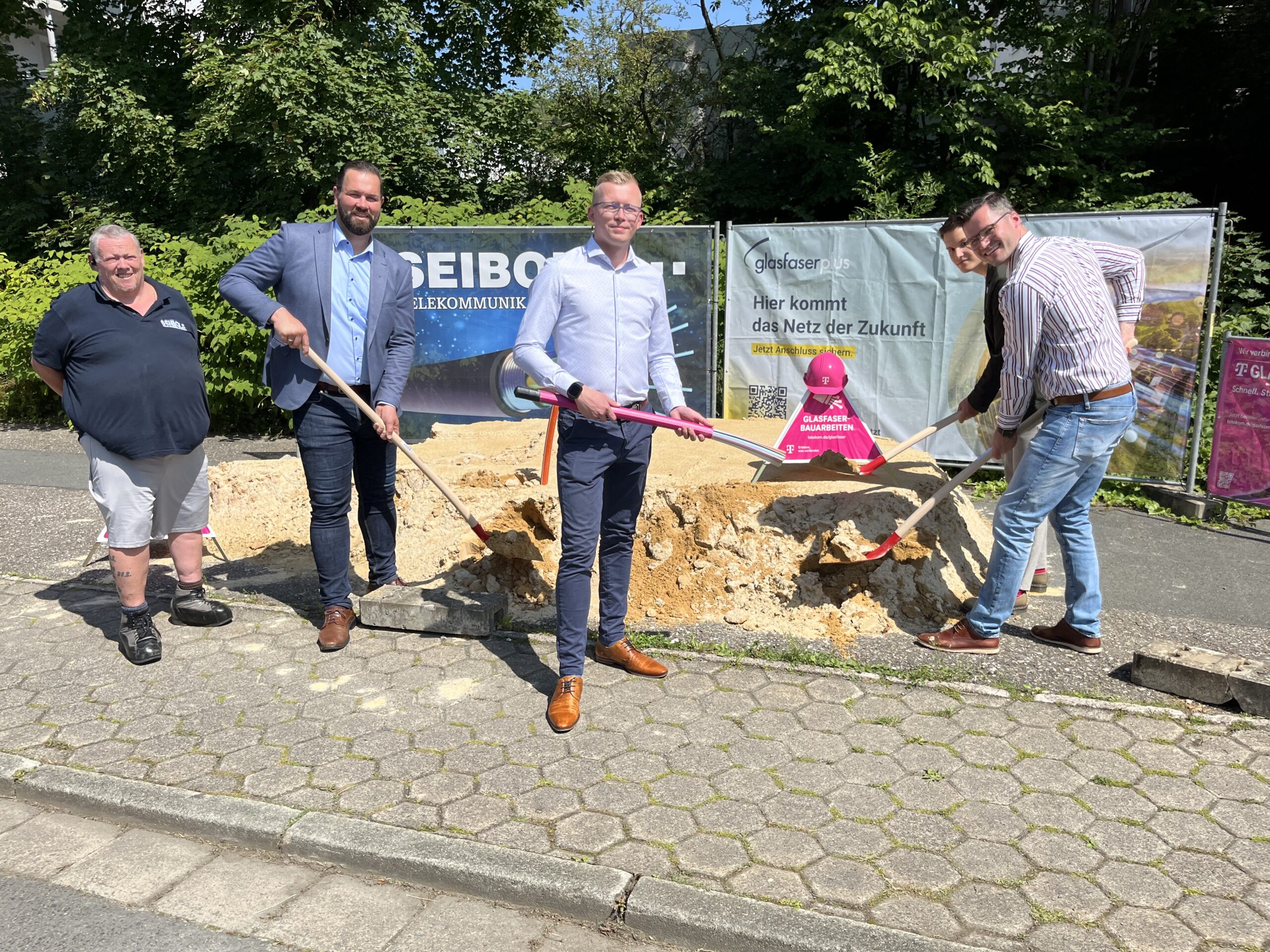 Vier Männer in Geschäftskleidung stehen auf einem Bürgersteig, halten Schaufeln in der Hand und posieren vor einer Baustelle mit Bannern und Erde, die den ersten Spatenstich für Glasfaser-Ausbau in Deutschland markiert.