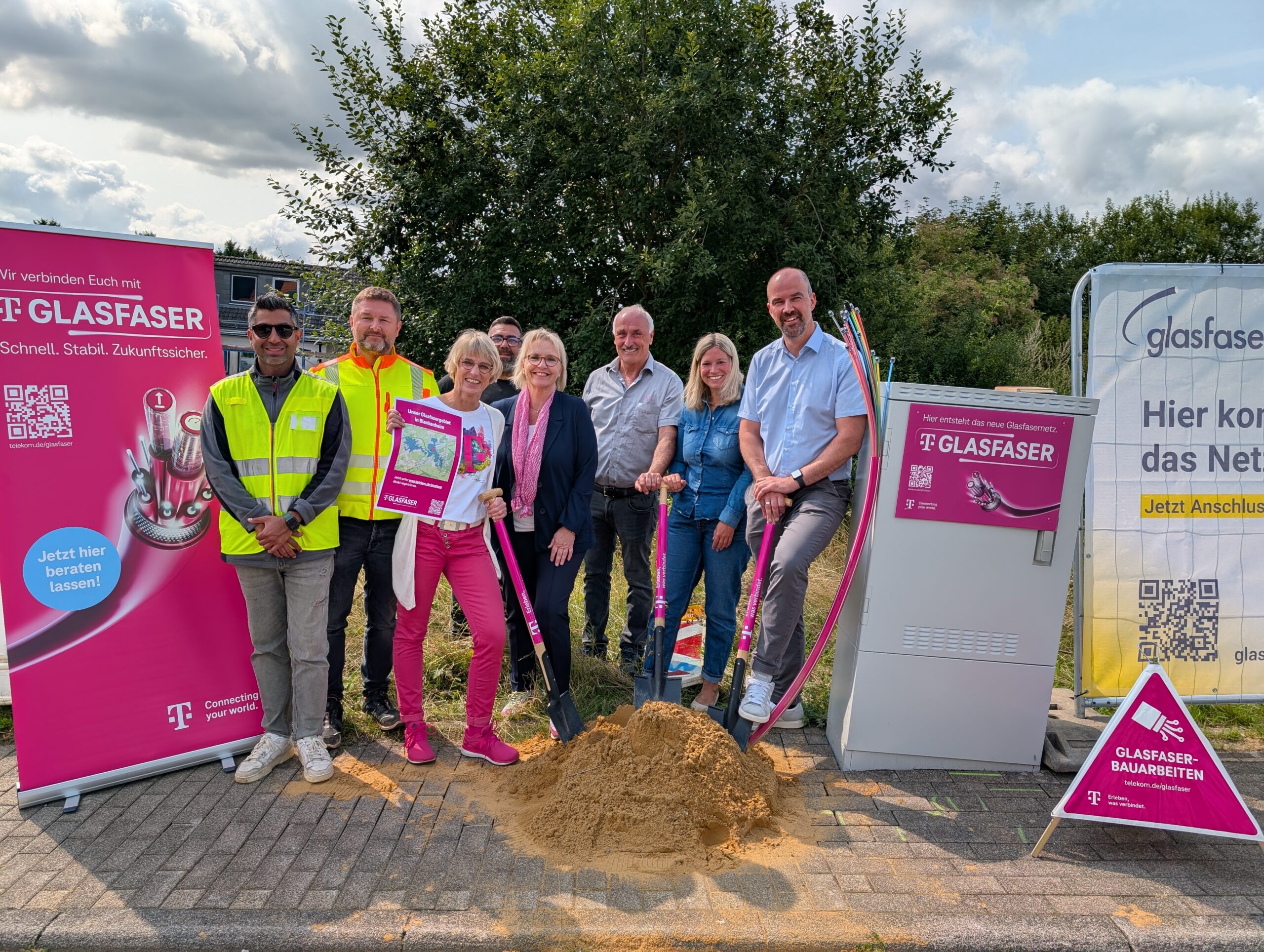 Eine Gruppe von Menschen posiert im Freien auf einer Glasfaser-Ausbau-Baustelle mit Schaufeln, neben Werbebannern und einem Telekommunikationsschrank.