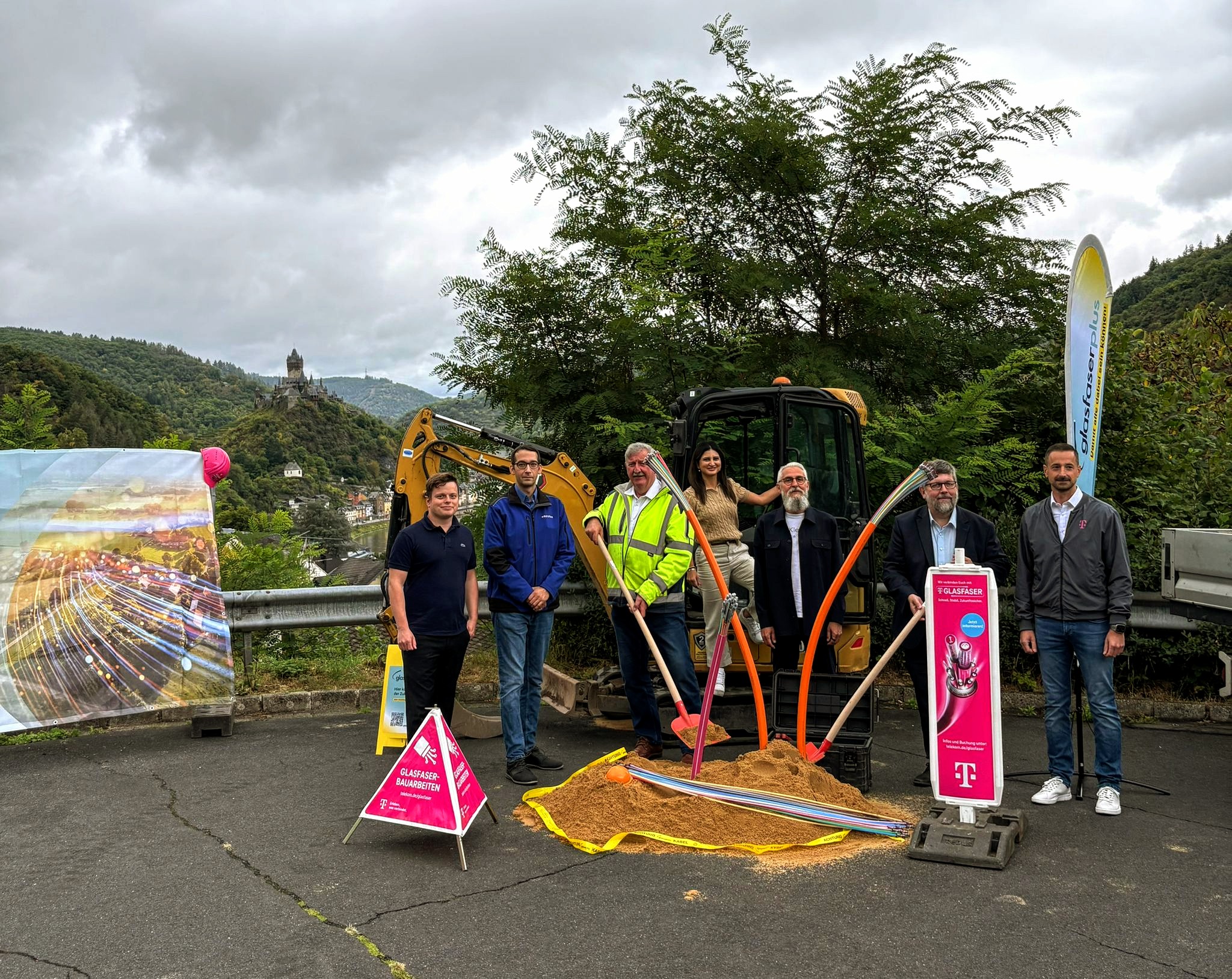 Eine Gruppe von Menschen steht beim Spatenstich von GlasfaserPlus neben einem kleinen Bagger und Telekom-Schildern, mit Schaufeln im Sand, im Freien in einer hügeligen Landschaft, um den Fortschritt beim Breitbandausbau zu markieren.