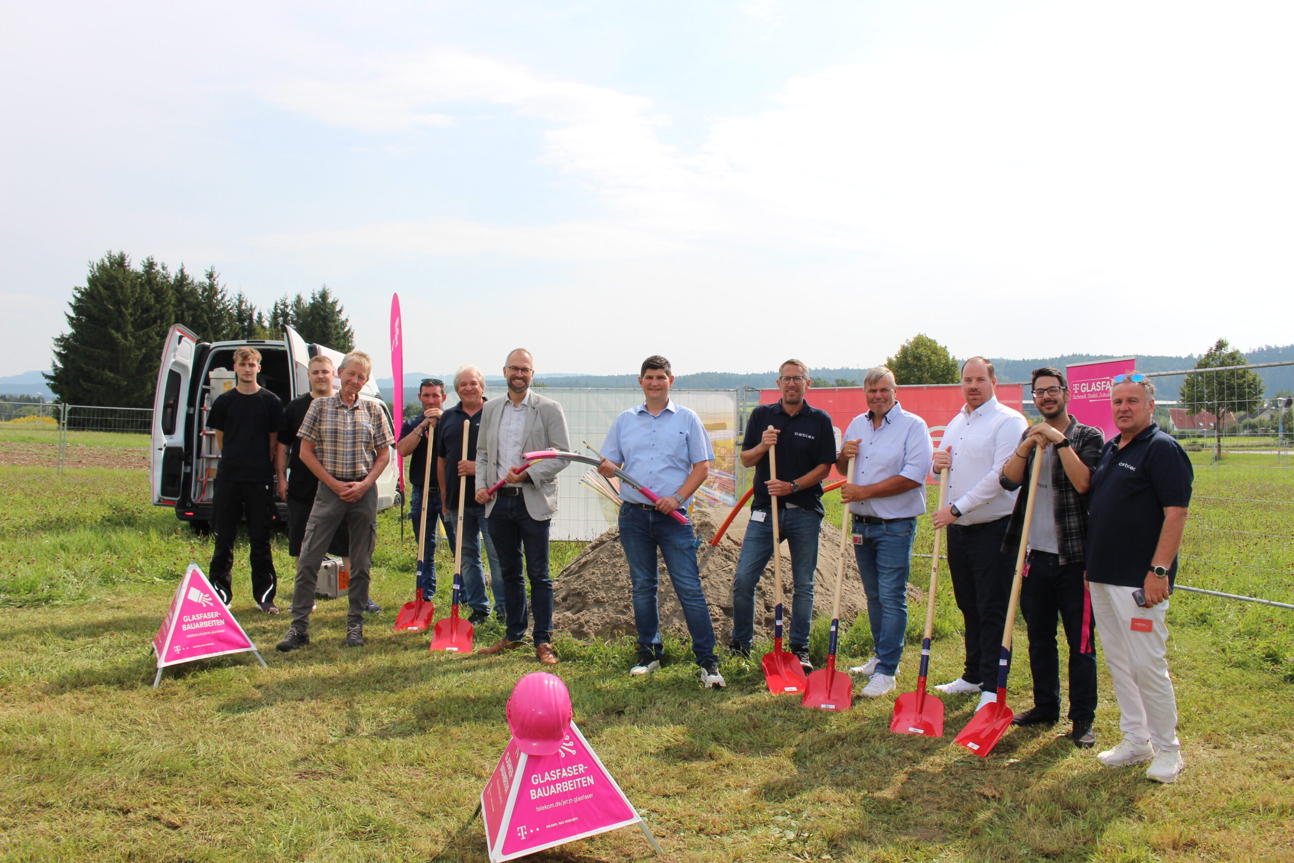 Eine Gruppe von Menschen steht im Freien mit Schaufeln in der Hand bei einer Spatenstichfeier für den Glasfaser-Ausbau in Deutschland, mit Bauschildern und Geräten auf einer Wiese.