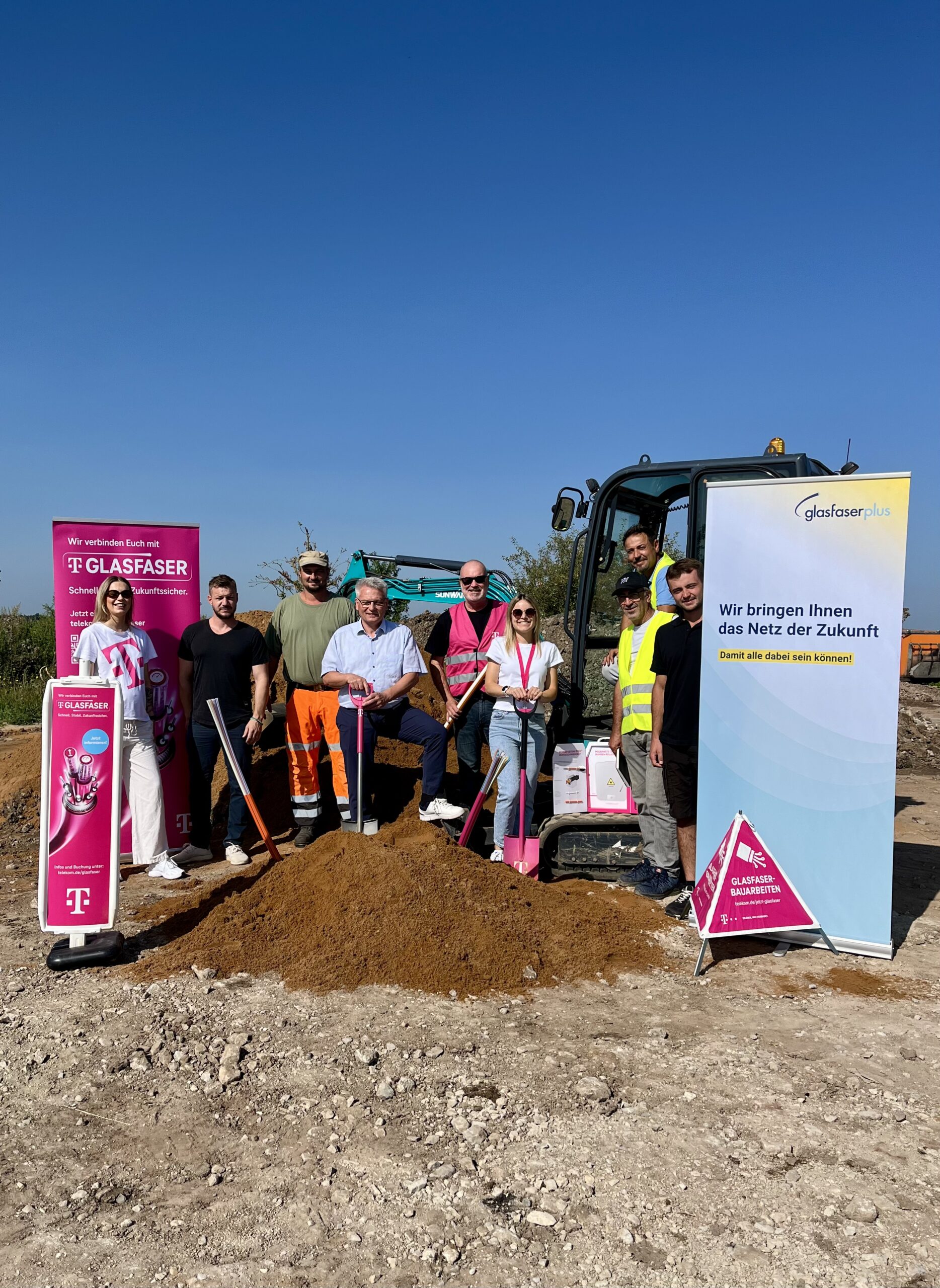 Eine Gruppe von Menschen steht mit Schaufeln auf der Baustelle von Glasfaser-Ausbau in Deutschland, vor Bannern und Baugeräten an einem sonnigen Tag.