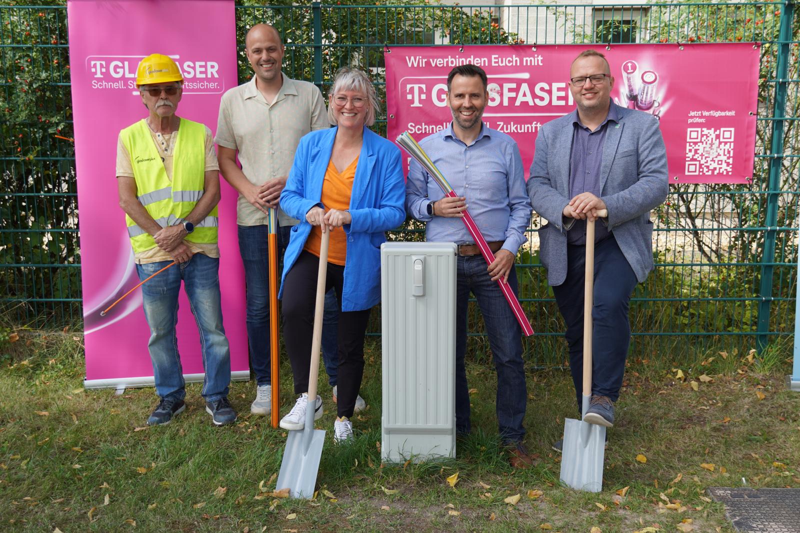 Fünf Personen stehen im Freien neben einem Glasfaserausbaukasten, halten Schaufeln und Kabel in der Hand und werben mit Bannern der Telekom für den Glasfaser-Ausbau in Deutschland und schnelles Internet.