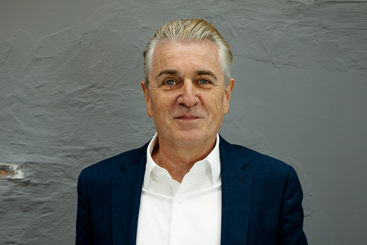 Ein Mann mittleren Alters mit grauen Haaren, der ein weißes Hemd und einen marineblauen Blazer trägt, steht vor einer strukturierten grauen Wand, blickt in die Kamera und lächelt leicht.