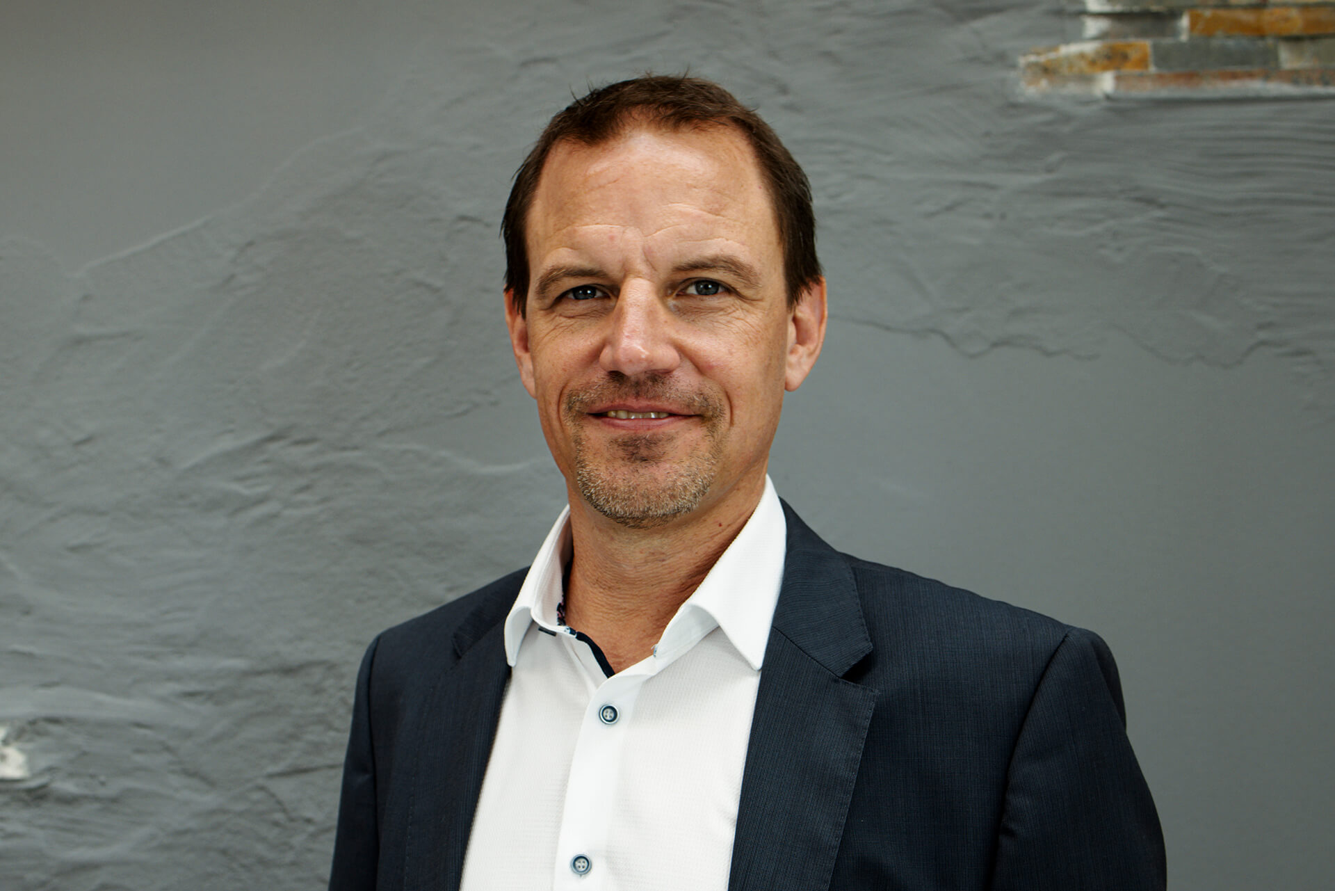 Ein Mann in einem dunklen Anzug und einem weißen Hemd steht vor einer strukturierten grauen Wand und blickt mit ausdruckslosem Gesicht in die Kamera.