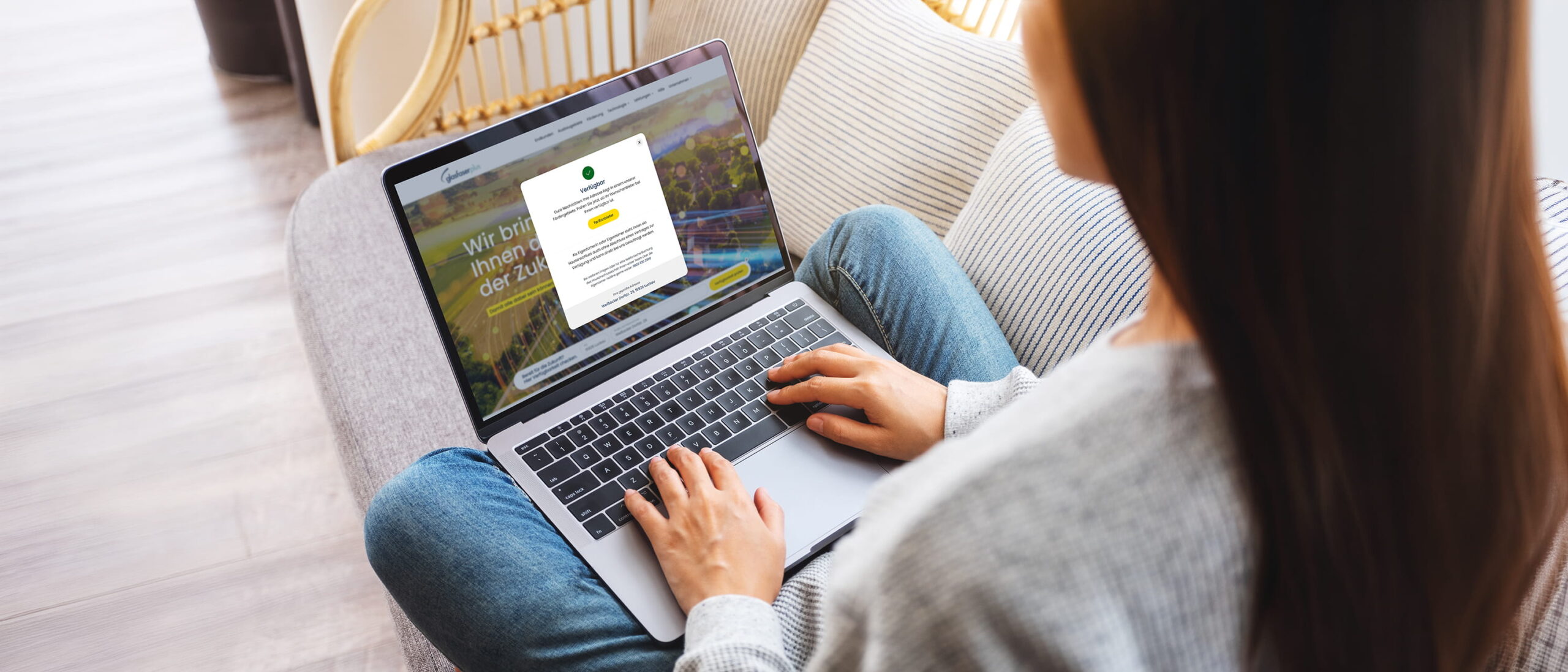Eine Person sitzt auf einem Sofa und nutzt einen Laptop, auf dessen Bildschirm eine Website mit deutschem Text sowie ein Online-Anmelde- oder Registrierungsformular geöffnet ist.