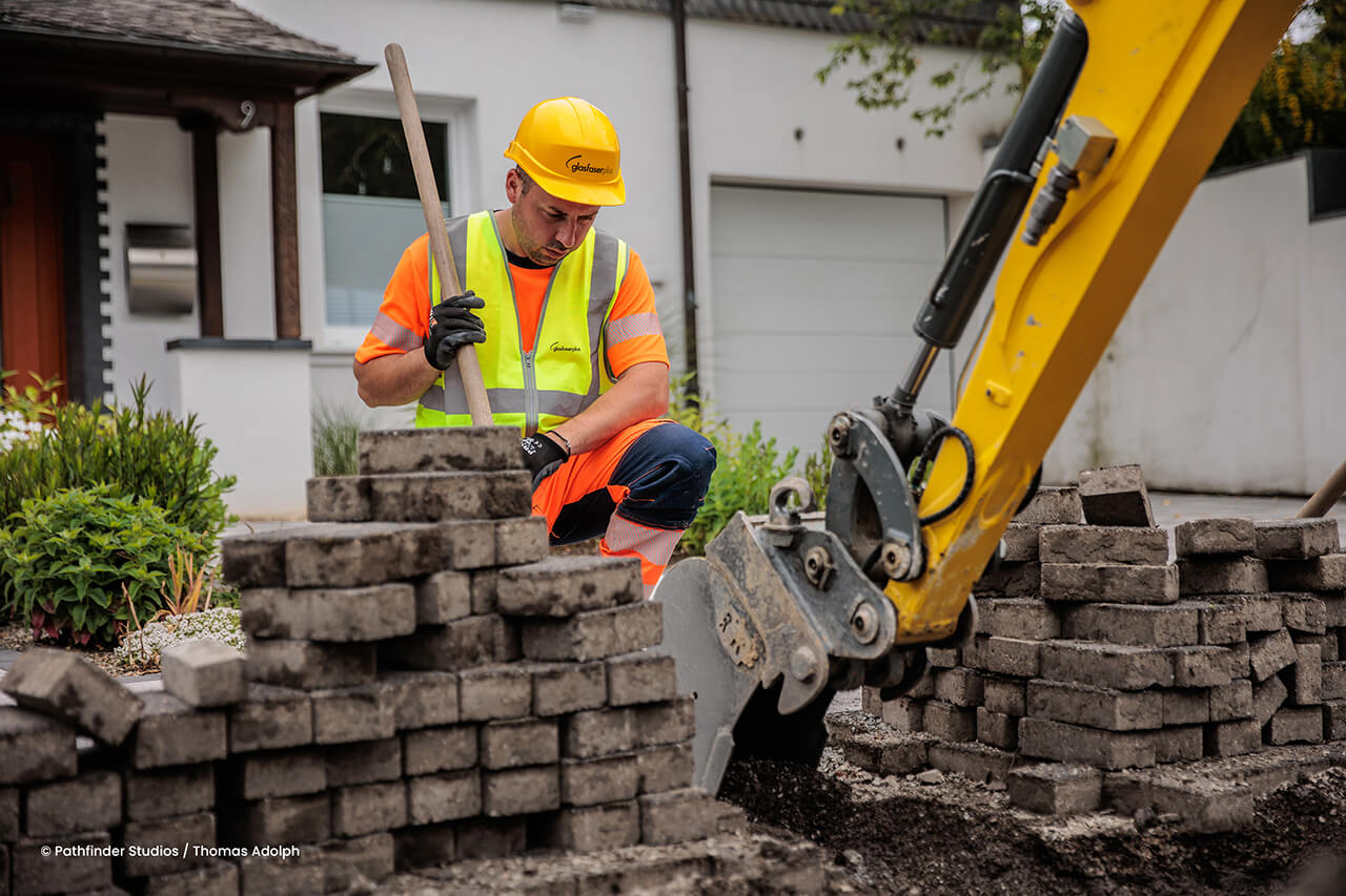 Ein Bauarbeiter mit gelbem Helm und gelber Weste arbeitet mit einer Schaufel neben einem Ziegelstapel und einem kleinen Bagger vor einem Wohnhaus.