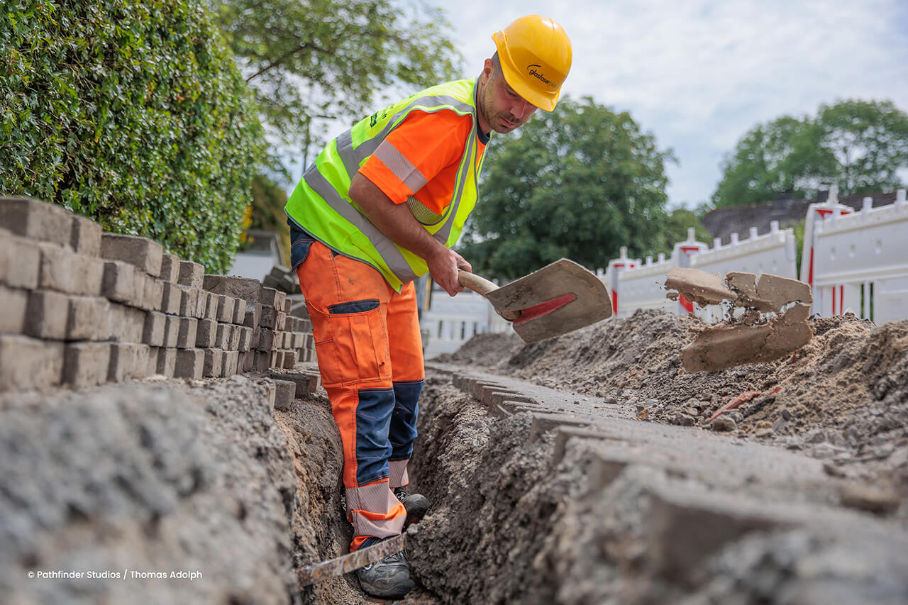 Ein Bauarbeiter mit gelbem Schutzhelm und reflektierender Schutzkleidung gräbt mit einer Schaufel einen Graben auf einer Baustelle im Freien, die mit Ziegelsteinen und Sicherheitsbarrieren eingefasst ist.