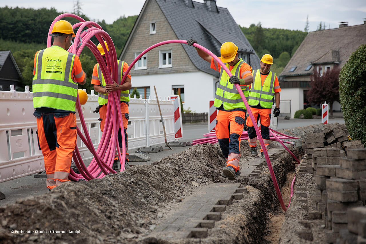 Vier Bauarbeiter in Schutzkleidung verlegen rosa Kabel in einem Graben in einer Wohnstraße; im Hintergrund sind Häuser und Absperrungen zu sehen.