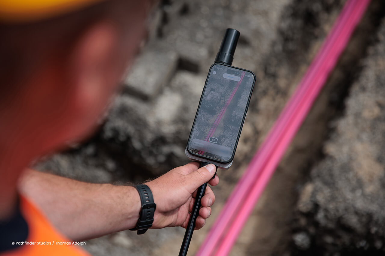 Eine Person hält ein Smartphone mit einem daran angebrachten Sensor in der Hand, auf dessen Bildschirm eine Karte über einer felsigen Ausgrabungsstätte zu sehen ist, auf der eine rosa Hilfslinie erkennbar ist.