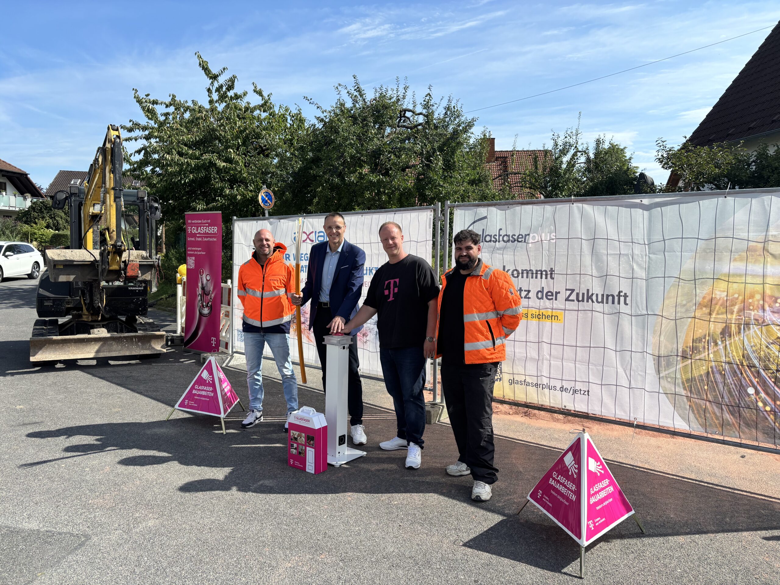 Vier Personen, darunter zwei in orangefarbenen Sicherheitswesten, stehen an einem sonnigen Tag auf einer Breitbandausbaustelle für die Glasfaserinstallation neben Baugeräten und Werbebannern.