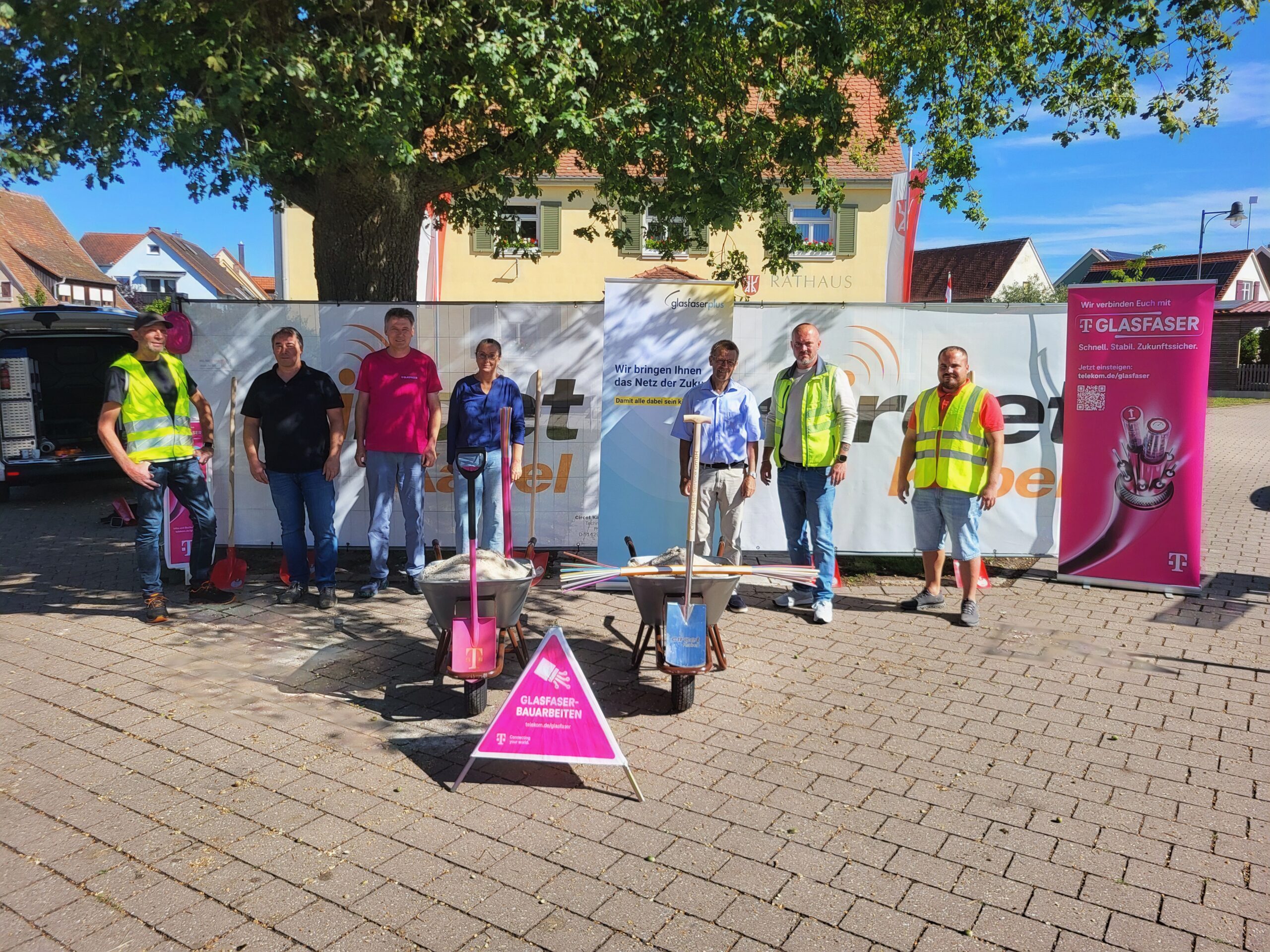 Sieben Personen stehen im Freien auf einer gepflasterten Fläche in der Nähe von Baumaschinen und Werbebannern für GlasfaserPlus, die auf den Glasfaser-Ausbau hinweisen. Im Hintergrund sind ein Baum und Gebäude zu sehen.