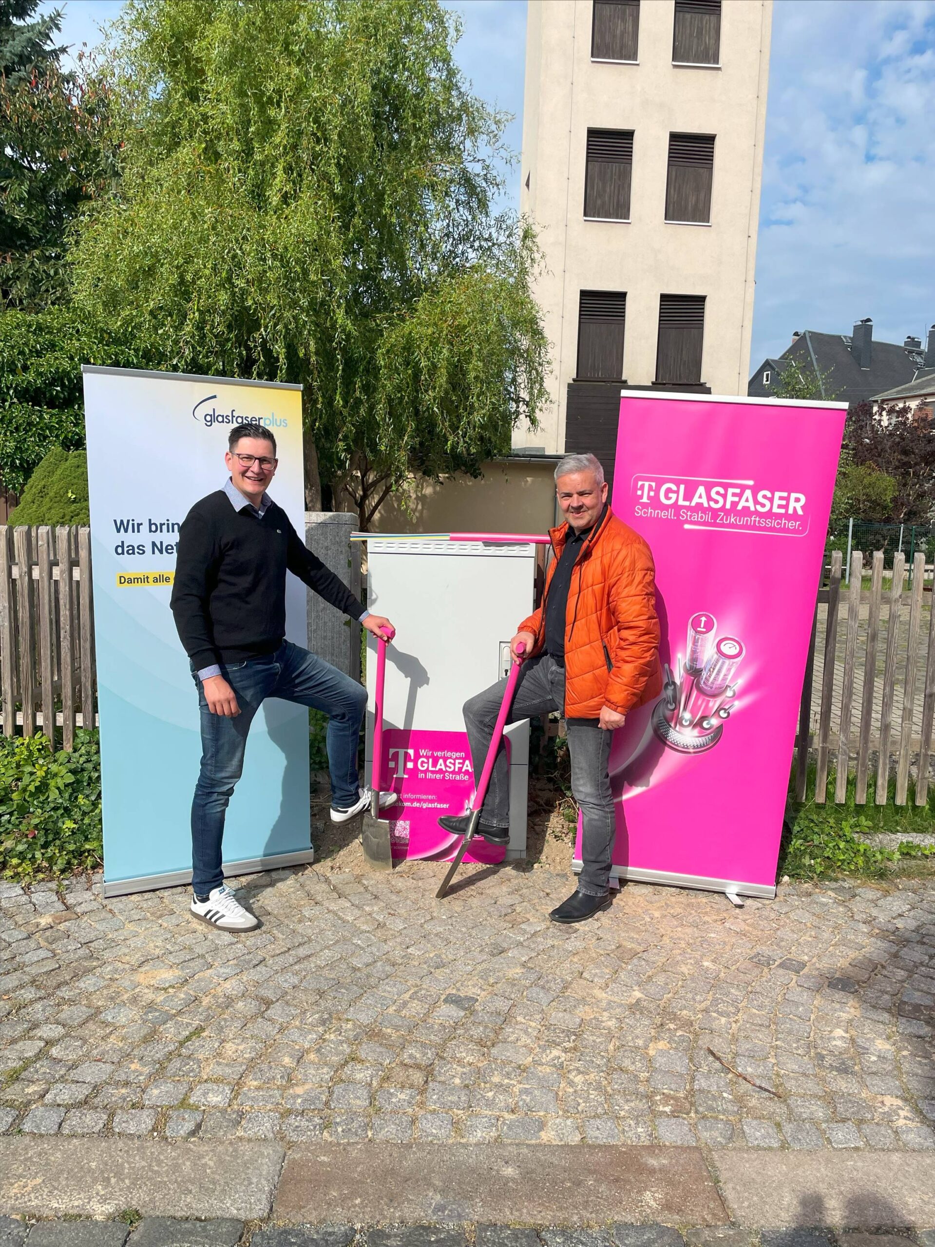 Zwei Männer posieren mit Werkzeugen neben einem Glasfaser-Internetschrank auf einem Gehweg. Im Hintergrund sind Werbebanner für GlasfaserPlus und Telekom Glasfaser zu sehen, die den Glasfaser-Ausbau in Deutschland präsentieren.