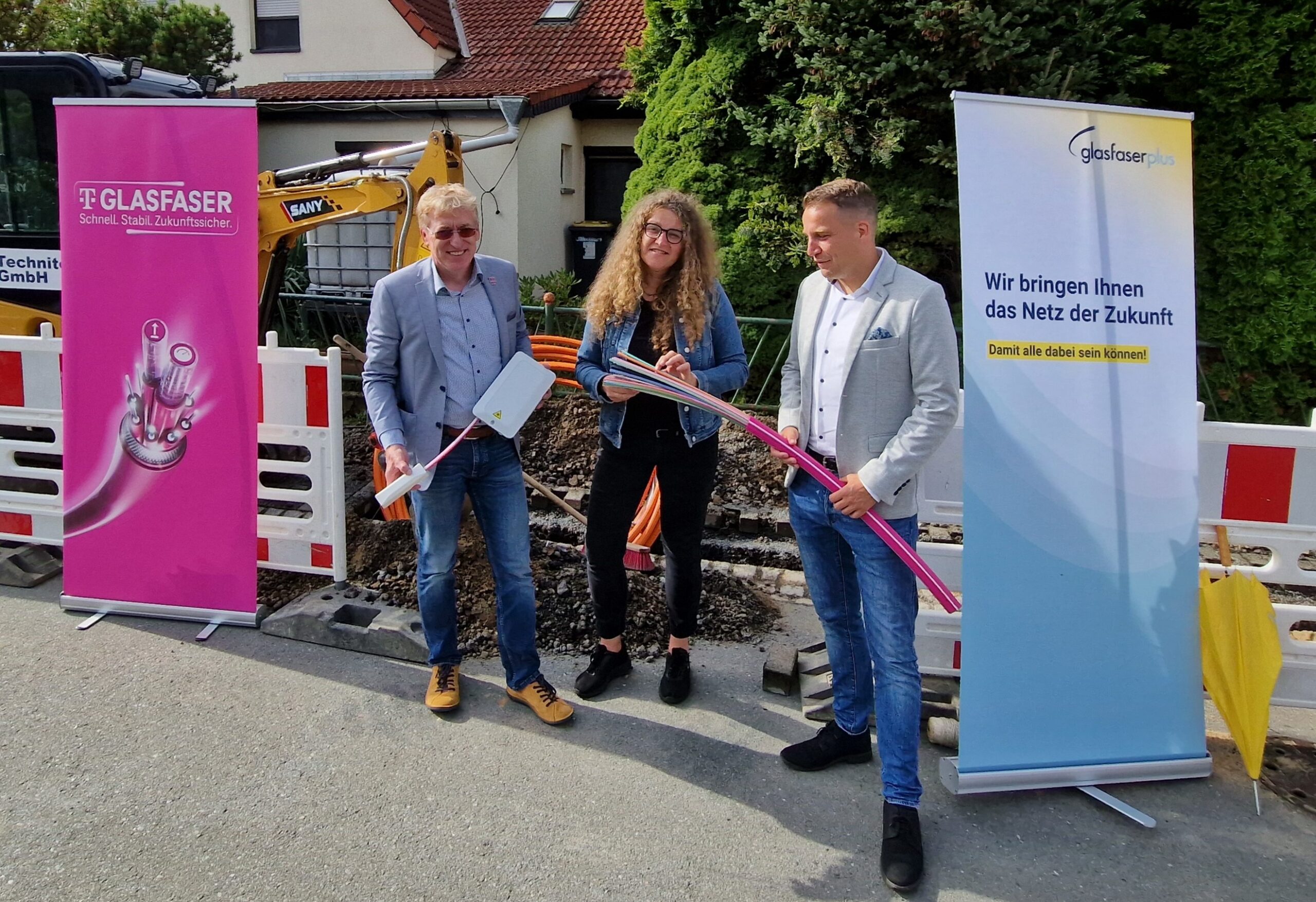 Drei Personen stehen an einer Baustelle und halten Glasfaserkabel. Im Hintergrund sind Werbebanner für den Glasfaser-Ausbau in Deutschland und den Breitbandausbau zu sehen.