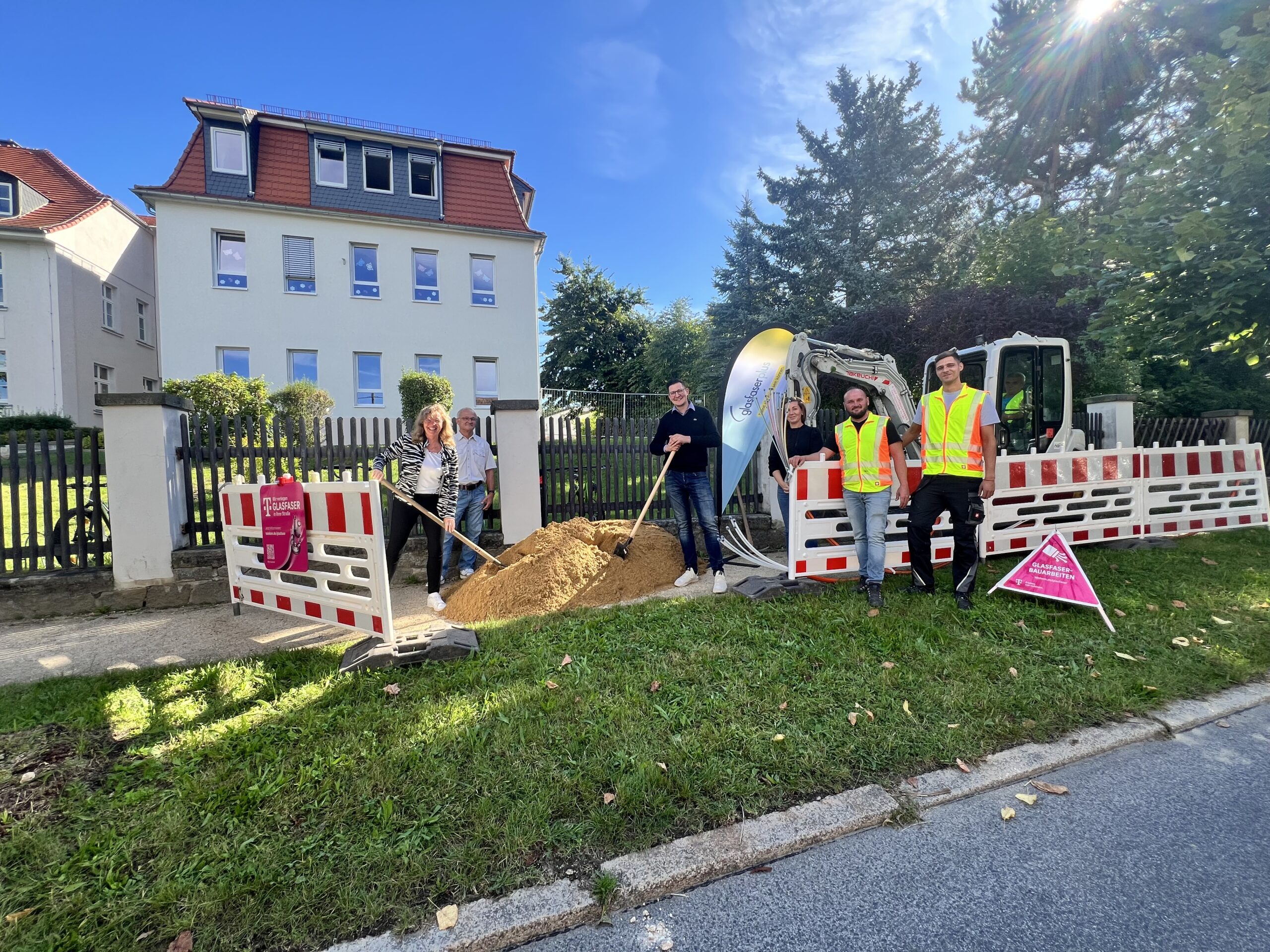 Vier Personen stehen an einer Baustelle mit kleinem Bagger und Sandhaufen, neben einer Wohnstraße hinter Schutzplanken, als GlasfaserPlus an einem sonnigen Tag am Breitbandausbau arbeitet.