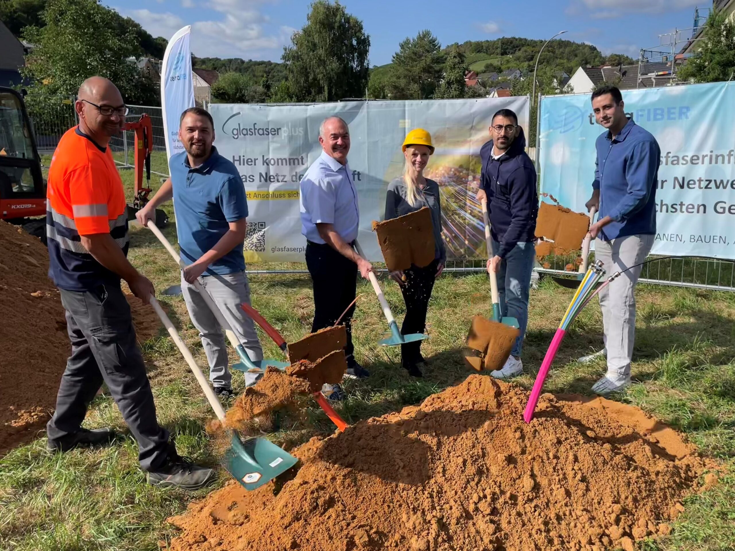 Sechs Personen stehen im Freien, halten Schaufeln in der Hand und posieren mit Erde bei einem Spatenstich für GlasfaserPlus, der den Glasfaser-Ausbau in Deutschland feiert. Im Hintergrund sind Baumaterialien und Transparente zu sehen.