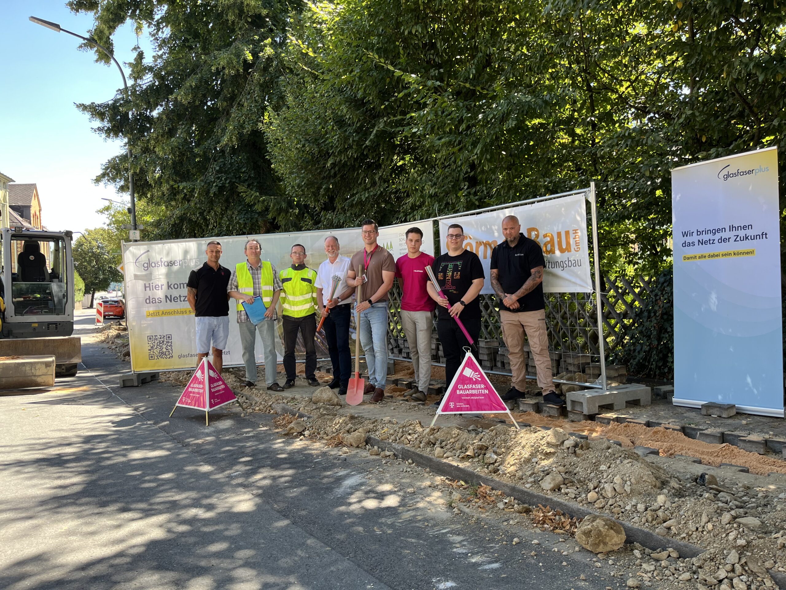 Eine Gruppe von Menschen steht auf einer Baustelle neben Schildern und Geräten, davor eine Schaufel und ein aufgegrabener Straßenabschnitt - die Szene verdeutlicht den Glasfaser-Ausbau in Deutschland.