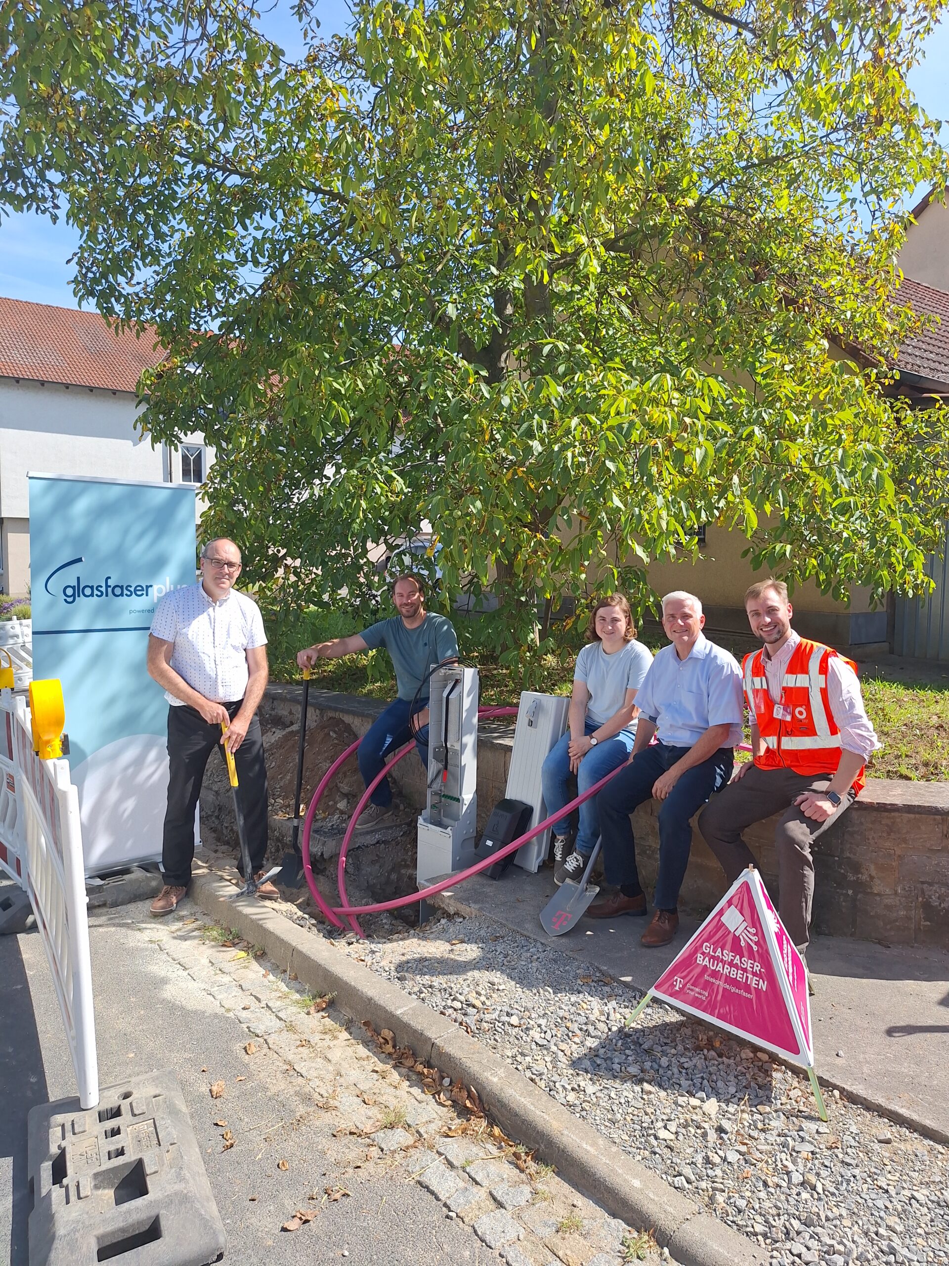 Fünf Personen, darunter eine in einer Sicherheitsweste, stehen neben Glasfaserkabeln und -geräten an einer sonnigen Straße, die auf den Glasfaser-Ausbau in Deutschland hinweist, mit einem Baum und einem Gebäude im Hintergrund.