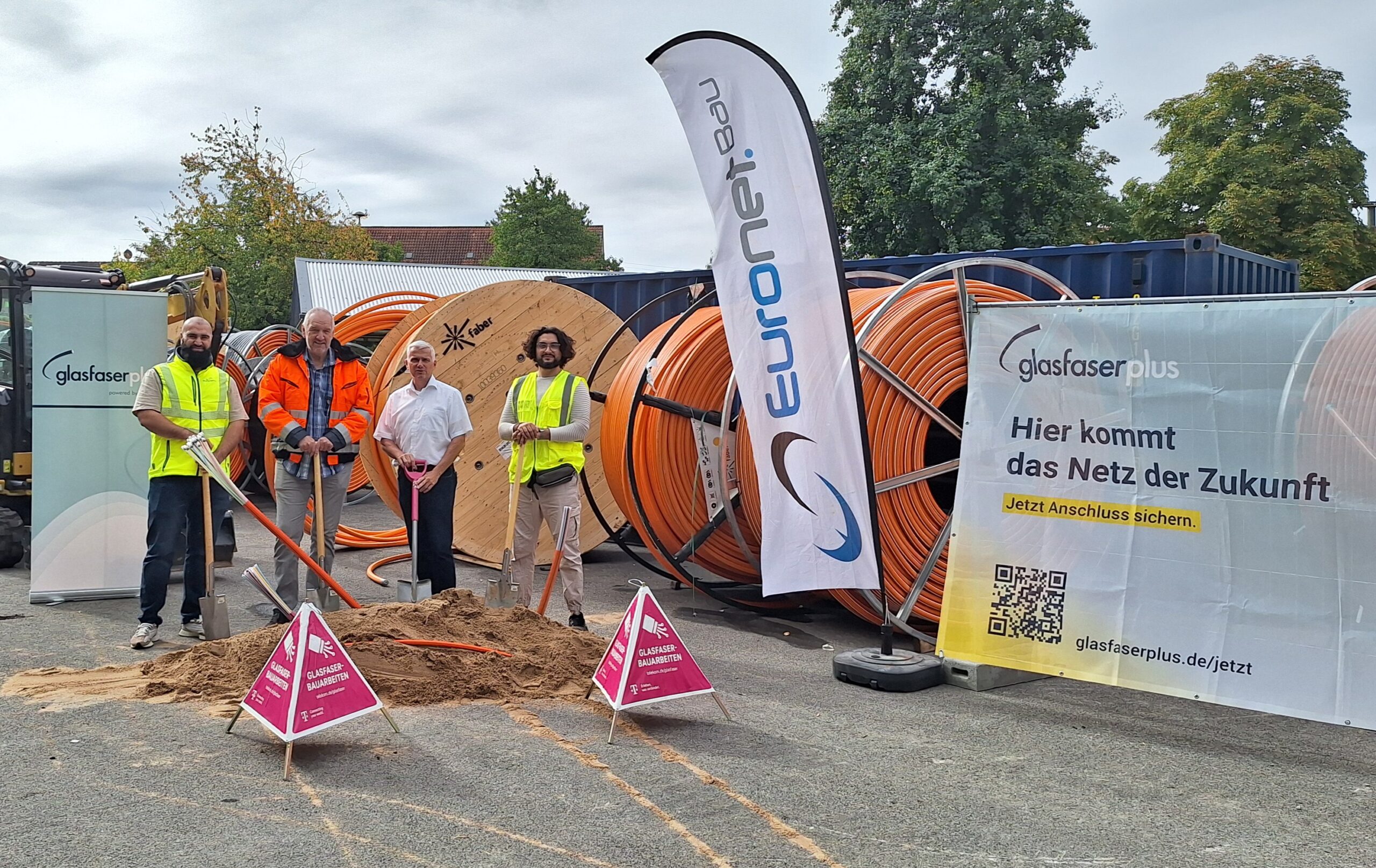 Vier Personen in Sicherheitswesten und Helmen stehen neben einem Sandhaufen und Glasfaserkabeln auf einer Baustelle mit Bannern für GlasfaserPlus, die auf den Fortschritt im Glasfaser-Ausbau hinweisen.