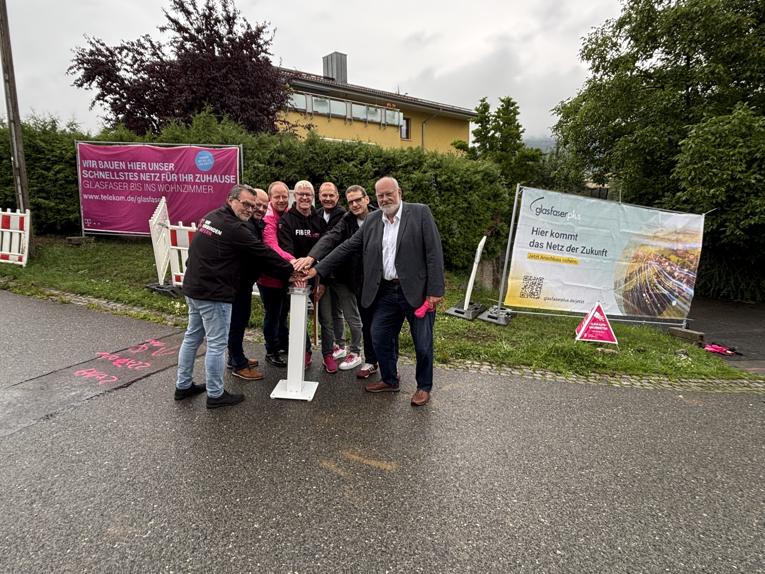Eine Gruppe von Menschen steht im Freien um einen weißen Pfosten herum und posiert für ein Foto. Im Hintergrund sind Bauschilder über Glasfaser-Ausbau in Deutschland zu sehen.