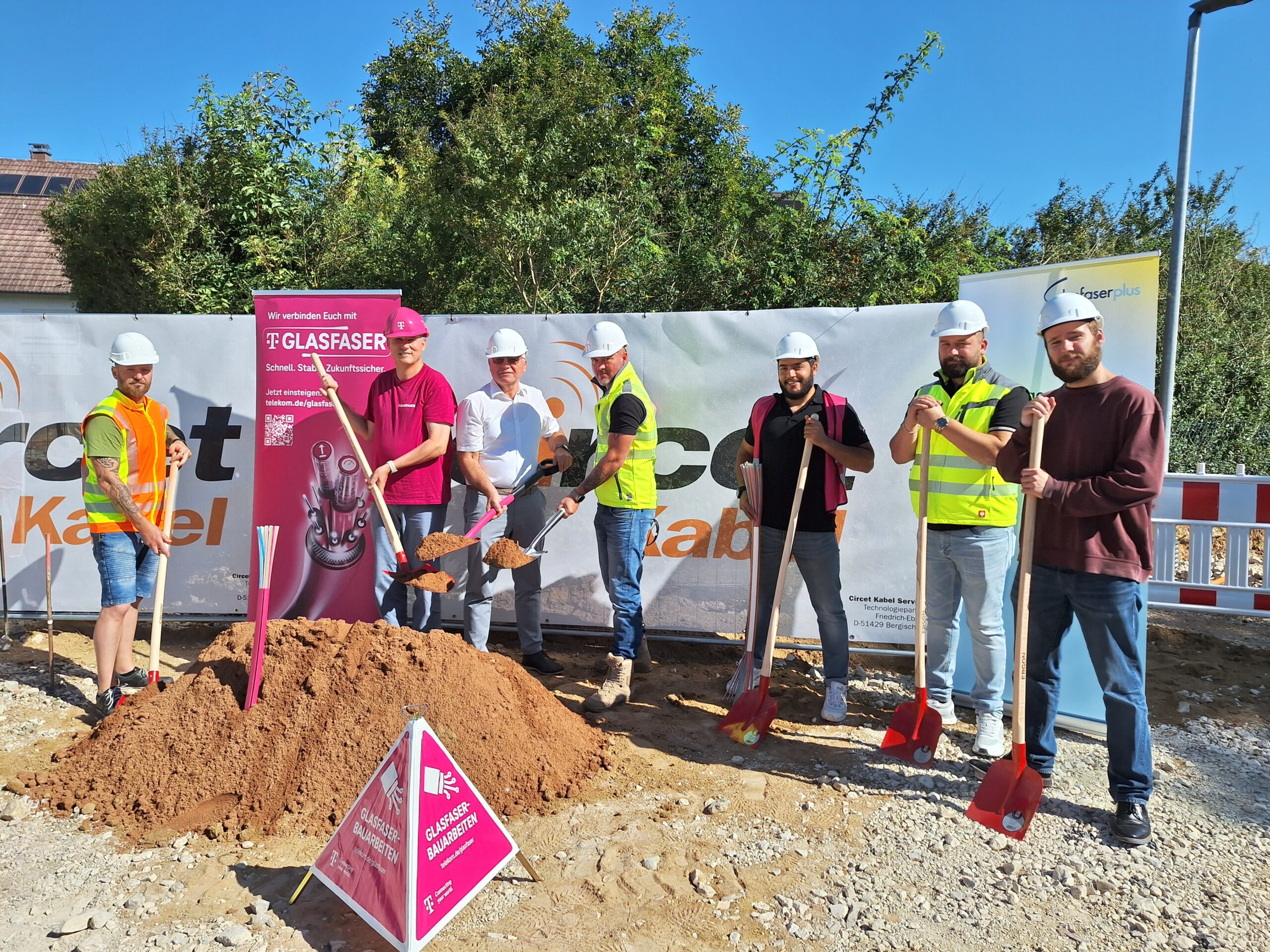 Sieben Personen in Baukleidung stehen mit Schaufeln an einer Baustelle, die den Startschuss für den Glasfaser-Ausbau in Deutschland gibt. Hinter ihnen sind Banner, ein Haufen Erde und Baumaschinen zu sehen.