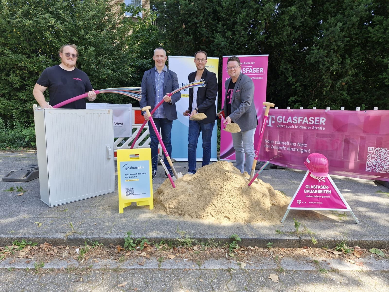 Vier Männer stehen auf einem Gehweg neben Baumaschinen, Glasfaserkabeln und einem Sandhaufen, feiern den Spatenstich für den Glasfaserausbau in Tönisvorst und werben für die Verlegung des neuen Glasfasernetzes.