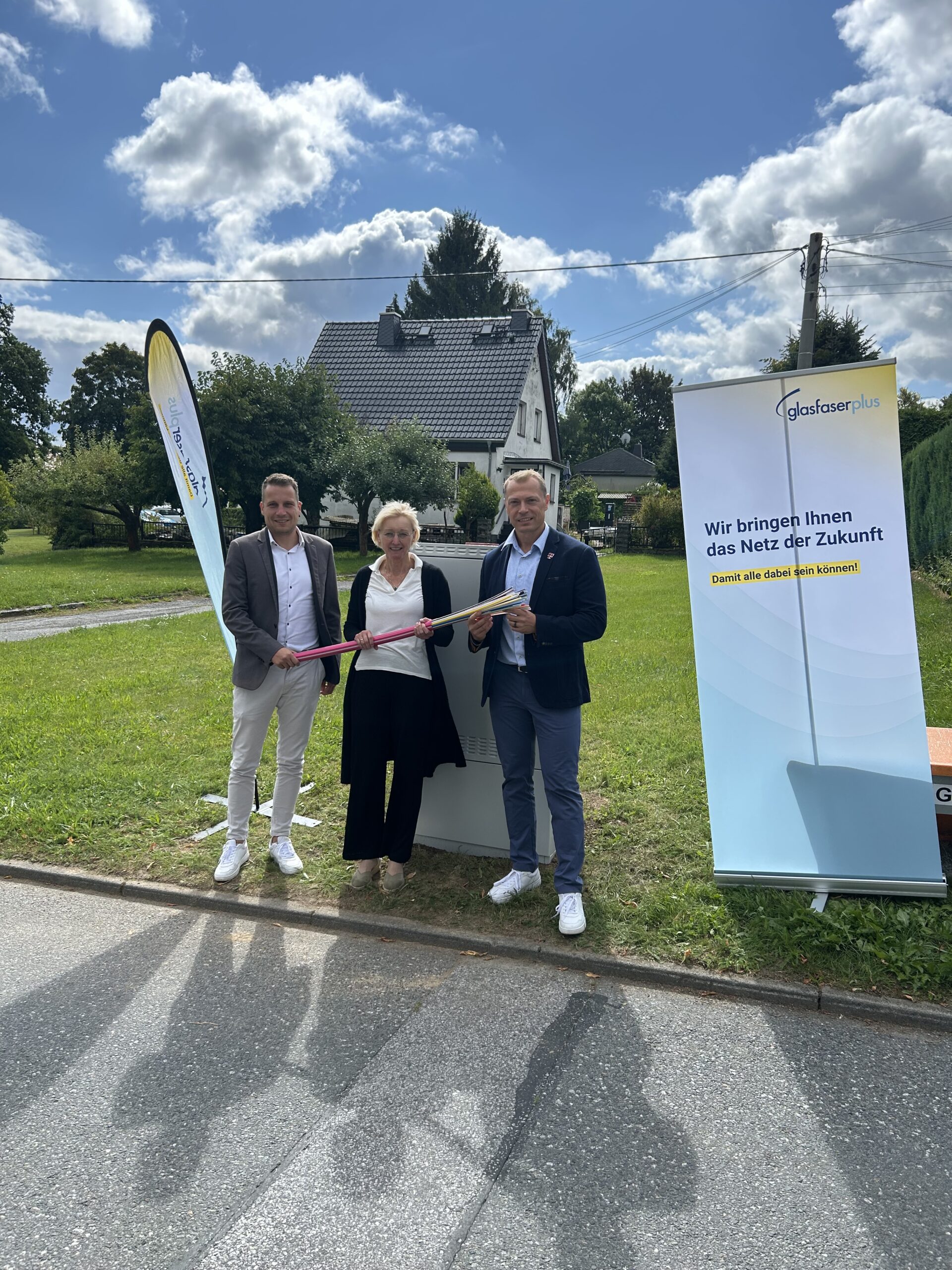Drei Personen stehen draußen an einer Straße und halten ein Band; Banner werben für Glasfaser-Ausbau und Hochgeschwindigkeits-Internet der Zukunft. Im Hintergrund sind ein Haus und Grünflächen zu sehen.