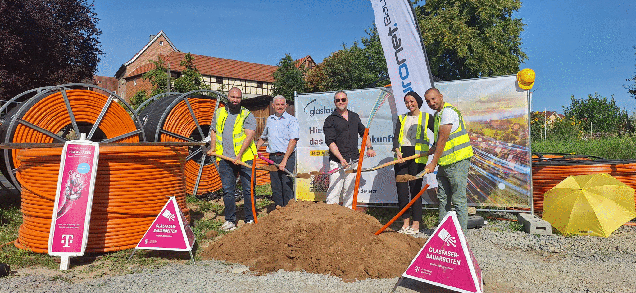 Fünf Personen in Sicherheitswesten stehen im Freien und halten Schaufeln bei einem Spatenstich für den Glasfaser-Ausbau in Deutschland. Im Hintergrund sind große orangefarbene Kabelrollen und ein Banner zu sehen.