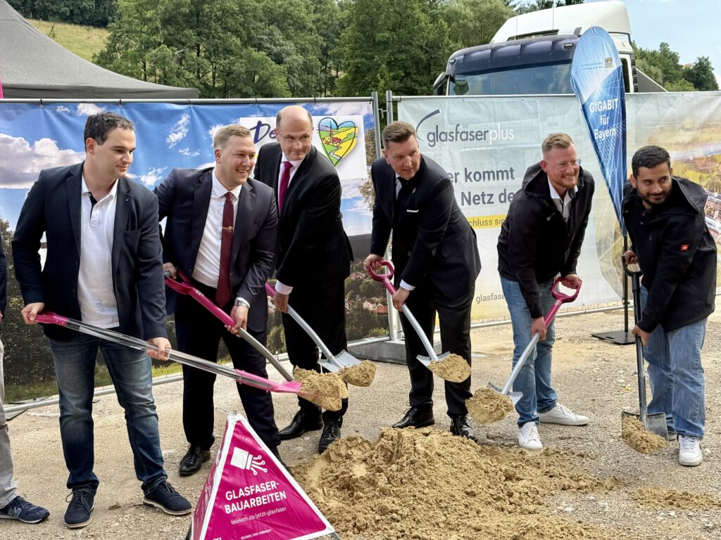 Fünf Männer in Anzügen und ein Mann in einem legeren Jackett nehmen am ersten Spatenstich für den Glasfaser-Ausbau in Deutschland teil. Sie halten Schaufeln in der Hand und posieren neben einem Haufen Erde auf der Baustelle.