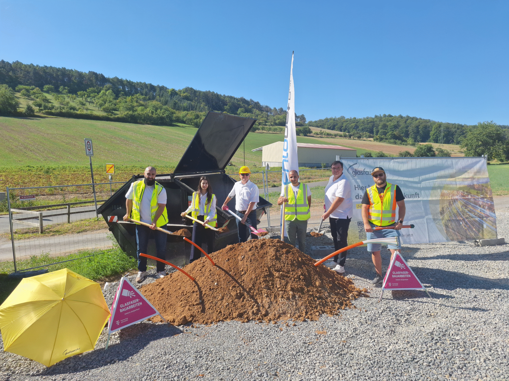 Fünf Personen in Sicherheitswesten und mit Schaufeln stehen an einem Erdhaufen auf einer ländlichen Baustelle, in der Nähe eines gelben Regenschirms und von Schildern, die den Fortschritt des Glasfaser-Ausbaus anzeigen.