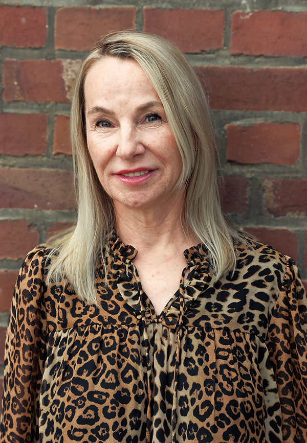 Eine Frau mit glattem blondem Haar, die eine Bluse mit Leopardenmuster trägt, steht vor einer Backsteinmauer, blickt in die Kamera und lächelt leicht.
